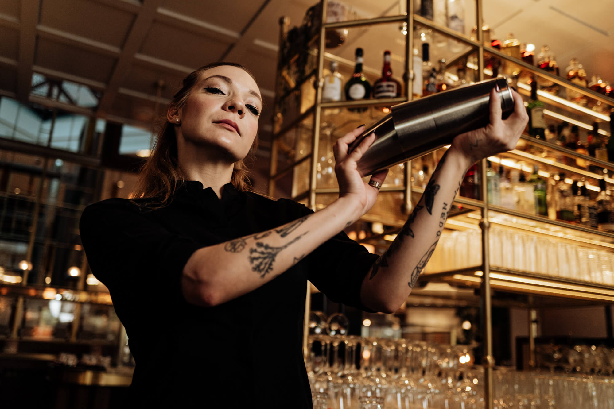 A bartender shakes a cocktail shaker as she stands in front of sparkling display of crystal glasses and liquor bottles