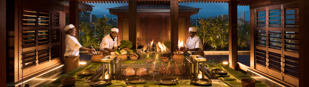 Ba đầu bếp trong một phòng ăn đông đúc của khách sạn Hilton ở Ấn Độ, mỗi người đứng xung quanh một chiếc bàn lớn hình vuông dành cho khách, trên đó có một xiên nướng gia cầm và một hố lửa hình tròn với những ngọn lửa màu cam bốc lên từ những tảng đá lớn nhẵn bóng.