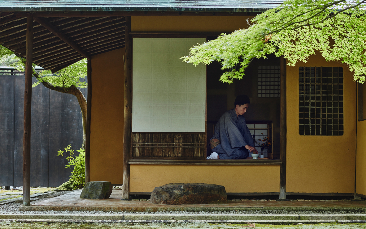 A man in a yukata grinds herbs with a mortal and pestle as he kneels in a traditional Japanese house