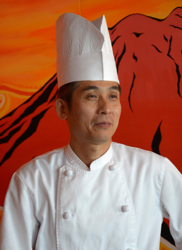 A Japanese chef wearing a tall white hat and white jacket, standing in front of a pop-art wall mural