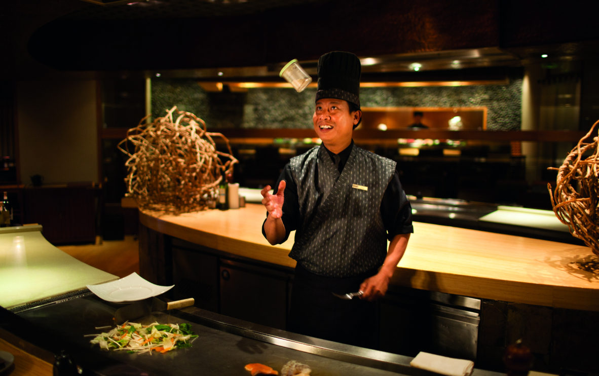 A Thai teppanyaki chef juggles a container of spice as he stands before a large steel grill