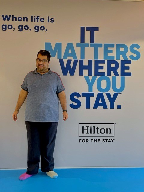 Person standing in front of a Hilton wall with text ‘It Matters Where You Stay’ and mismatched blue and yellow socks.