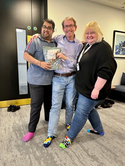 Three people standing together indoors wearing colorful patterned socks; one holds a book. Office chairs and artwork in background.