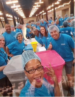 A large group of people wearing matching blue shirts and hairnets gathered in a spacious, well-lit room with long tables. Several colorful plastic bins, including pink and gray ones, are in the foreground, and some participants are giving thumbs-up gestures. The setting appears to be an organized volunteer or team-building event.